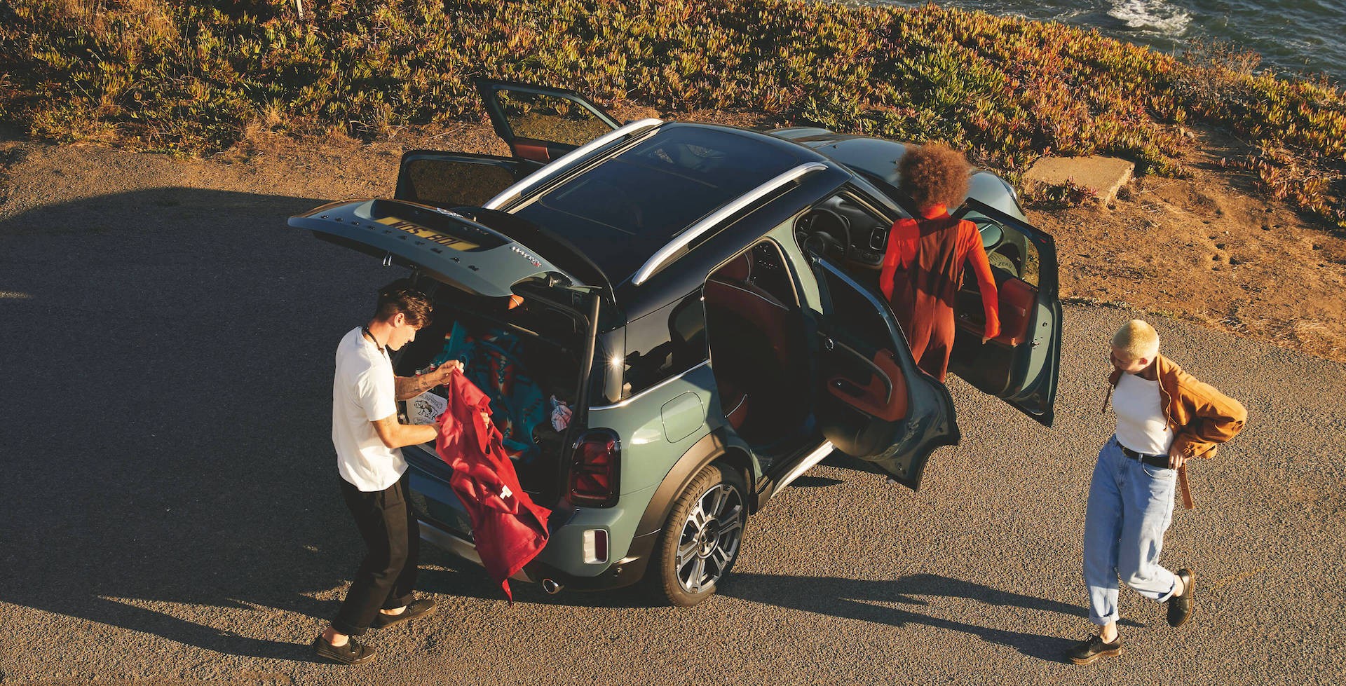 Three friends unpacking a green MINI Countryman parked near a coastal road at sunset. The car’s rear hatch and doors are open as one person retrieves clothes from the trunk.